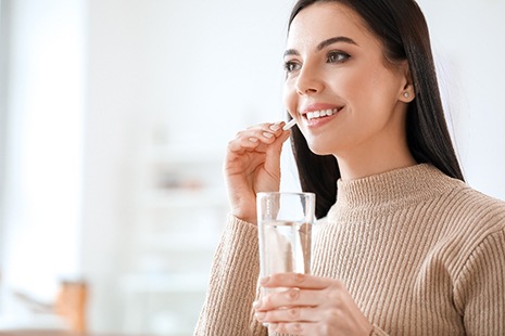 Woman in beige sweater taking a pill with a glass of water
