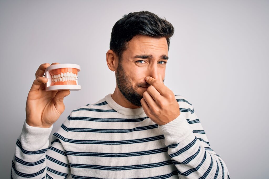 Man in striped sweater holding artificial teeth in one hand and plugging his nose with the other